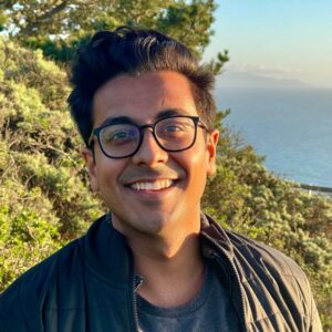 Manu Meel is smiling at the camera against a backdrop of trees and water. He is wearing black glasses, a dark jacket, and blue shirt.